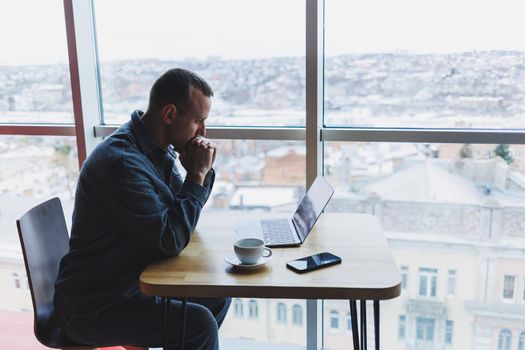 A young successful businessman in a suit drinks coffee and works in a cafe on wireless free internet on a laptop, sitting alone at a table in a cafe.