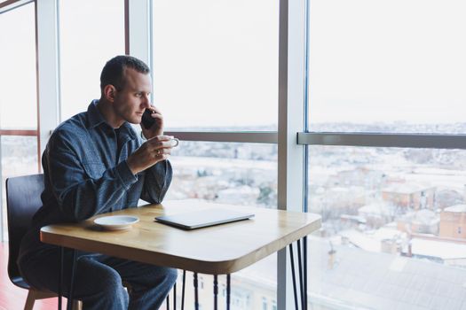 Happy man with coffee calling via smartphone while remote working laptop at work desk, cheerful freelancer enjoying coffee break while working remotely