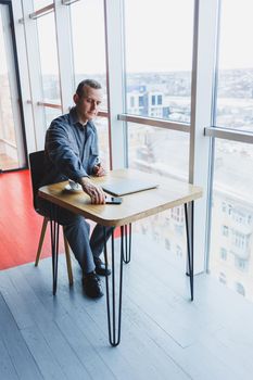 Happy man with coffee calling via smartphone while remote working laptop at work desk, cheerful freelancer enjoying coffee break while working remotely