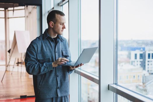 Portrait of a smart intelligent male manager, he is holding a laptop for research in the office, a business man in elegant clothes playing on the touchpad. Standing in the office by a large window