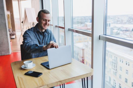 Successful happy business man is sitting at a table in a cafe, holding a cup of coffee and using a laptop.