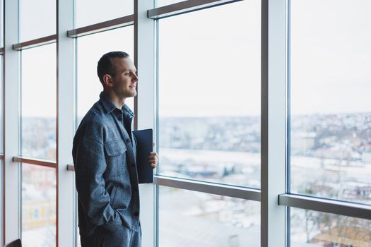 Portrait of a smart intelligent male manager, he is holding a laptop for research in the office, a business man in elegant clothes playing on the touchpad. Standing in the office by a large window