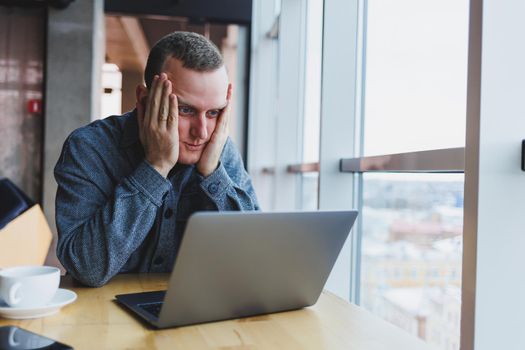 A young handsome man sits at a table and looks with surprise at the screen of his laptop, remote work