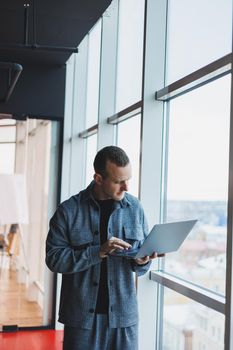 Portrait of a smart intelligent male manager, he is holding a laptop for research in the office, a business man in elegant clothes playing on the touchpad. Standing in the office by a large window