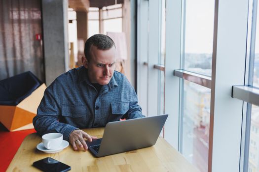 Successful happy business man is sitting at a table in a cafe, holding a cup of coffee and using a laptop.