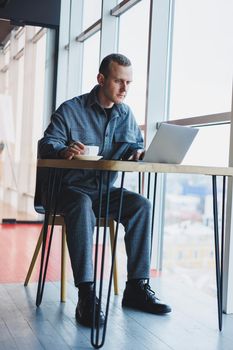 A young successful businessman in a suit drinks coffee and works in a cafe on wireless free internet on a laptop, sitting alone at a table in a cafe.