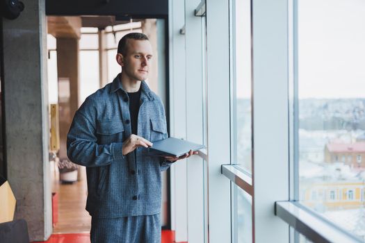 Portrait of a smart intelligent male manager, he is holding a laptop for research in the office, a business man in elegant clothes playing on the touchpad. Standing in the office by a large window