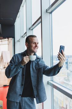 European male freelancer in checking notification on smartphone about money transactions, doing business online, working remotely while in cafe and drinking coffee