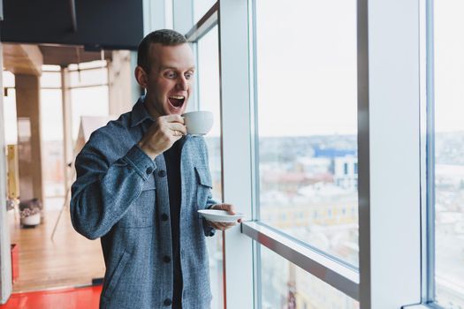 A young man of European appearance in a casual suit drinks coffee and looks out the window