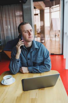 Smiling male entrepreneur in casual wear sitting at table with laptop and talking on smartphone in modern workspace with large window during day