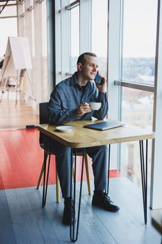 Cheerful IT professional with laptop talking on smartphone to discuss web programming, happy businessman in cafe drinking coffee. freelancer