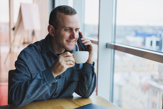 European male freelancer in checking notification on smartphone about money transactions, doing business online, working remotely while in cafe and drinking coffee