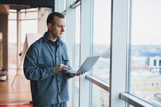 Portrait of a smart intelligent male manager, he is holding a laptop for research in the office, a business man in elegant clothes playing on the touchpad. Standing in the office by a large window