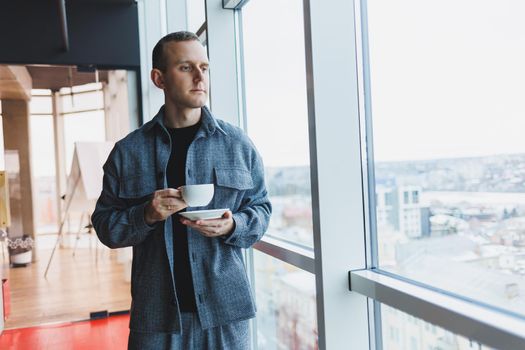 A young man of European appearance in a casual suit drinks coffee and looks out the window