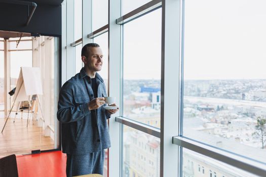 A young man of European appearance in a casual suit drinks coffee and looks out the window