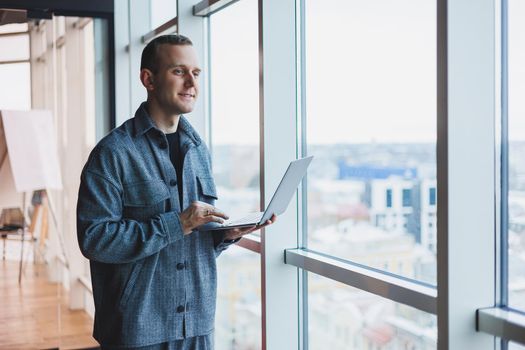 Portrait of a smart intelligent male manager, he is holding a laptop for research in the office, a business man in elegant clothes playing on the touchpad. Standing in the office by a large window