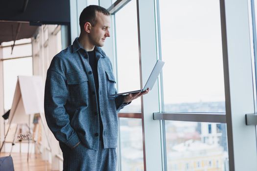 Portrait of a smart intelligent male manager, he is holding a laptop for research in the office, a business man in elegant clothes playing on the touchpad. Standing in the office by a large window
