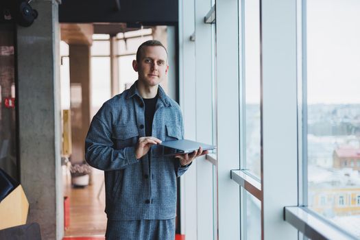 Portrait of a smart intelligent male manager, he is holding a laptop for research in the office, a business man in elegant clothes playing on the touchpad. Standing in the office by a large window