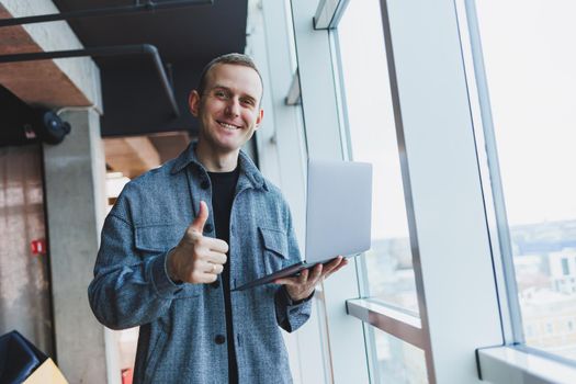 Portrait of a smart intelligent male manager, he is holding a laptop for research in the office, a business man in elegant clothes playing on the touchpad. Standing in the office by a large window