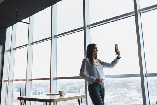 Young businesswoman wearing glasses and holding a phone. In an office or business center against the background of large windows of a skyscraper. Work and career, concept