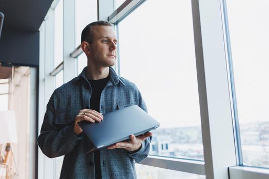Portrait of a smart intelligent male manager, he is holding a laptop for research in the office, a business man in elegant clothes playing on the touchpad. Standing in the office by a large window