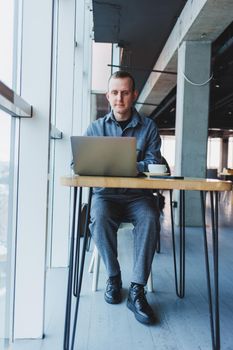 A young successful businessman in a suit drinks coffee and works in a cafe on wireless free internet on a laptop, sitting alone at a table in a cafe.