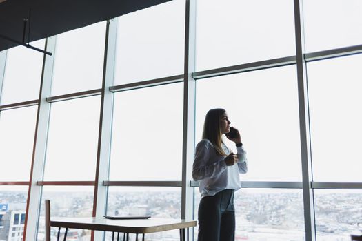 Young businesswoman wearing glasses and holding a phone. In an office or business center against the background of large windows of a skyscraper. Work and career, concept