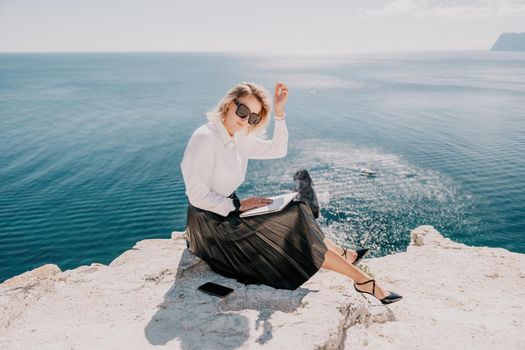 Successful business woman in yellow hat working on laptop by the sea. Pretty lady typing on computer at summer day outdoors. Freelance, travel and holidays concept.