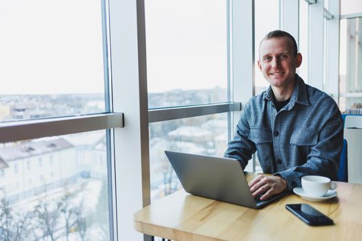 Successful happy business man is sitting at a table in a cafe, holding a cup of coffee and using a laptop.