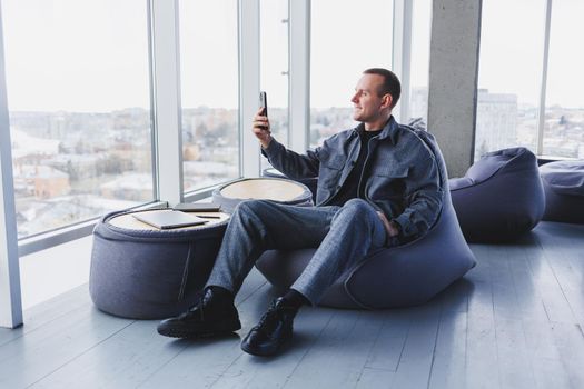 Young male student in a casual suit resting in a comfortable pouf while studying, an attractive freelancer in a suit works remotely