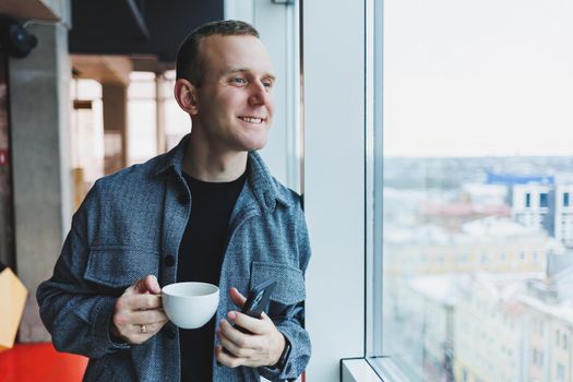A young man of European appearance in a casual suit drinks coffee and looks out the window