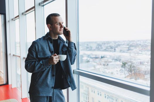 A handsome man of European appearance in a casual suit drinks coffee, talks on the phone and looks out the window