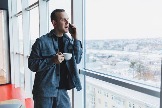 A handsome man of European appearance in a casual suit drinks coffee, talks on the phone and looks out the window