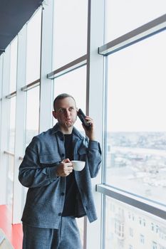 A handsome man of European appearance in a casual suit drinks coffee, talks on the phone and looks out the window