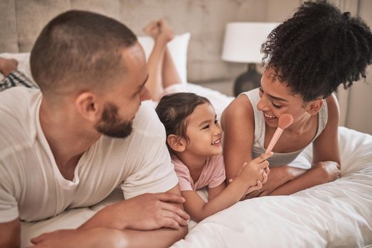 Cosmetics, relax and happy family on a bed with the child having fun with a makeup brush, laughing and playing. Mother, father and girl enjoy quality time together with beauty products in the morning.