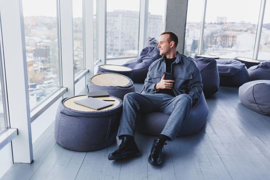 Young male student in a casual suit resting in a comfortable pouf while studying, an attractive freelancer in a suit works remotely