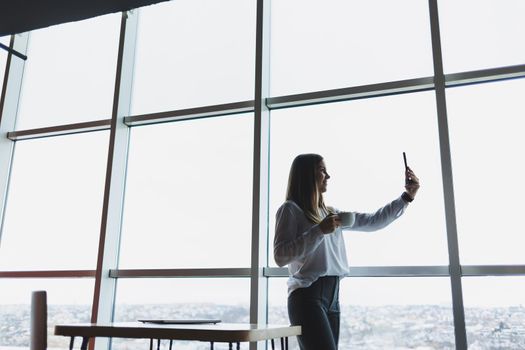 Young businesswoman wearing glasses and holding a phone. In an office or business center against the background of large windows of a skyscraper. Work and career, concept