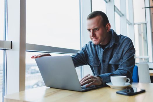 Successful happy business man is sitting at a table in a cafe, holding a cup of coffee and using a laptop.