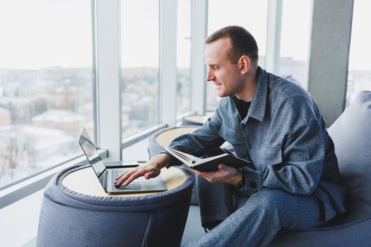 A young male student in a casual suit resting in a comfortable pouf while studying makes notes in a notebook, an attractive freelancer in a suit works remotely