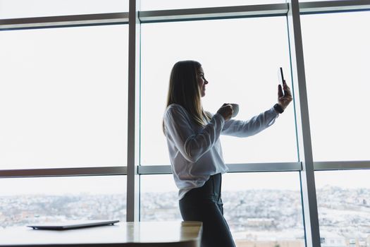 Young businesswoman wearing glasses and holding a phone. In an office or business center against the background of large windows of a skyscraper. Work and career, concept