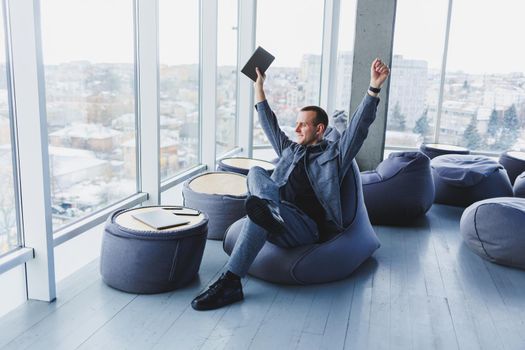 A young male student in a casual suit resting in a comfortable pouf while studying makes notes in a notebook, an attractive freelancer in a suit works remotely