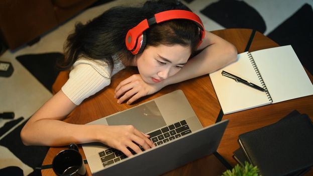 Overhead view of woman wearing headphone lying on wooden table and looking at laptop screen.