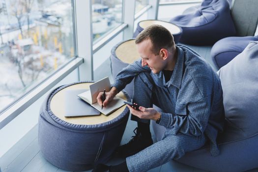 A handsome European young businessman in a coffee shop sits with a mobile phone and makes notes in a notebook. Wearing an elegant casual suit and sitting with a laptop in a public cafe