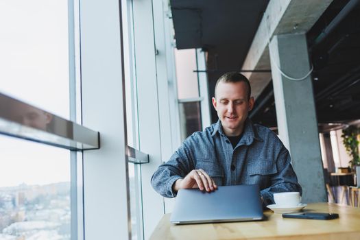 A young successful businessman in a suit drinks coffee and works in a cafe on wireless free internet on a laptop, sitting alone at a table in a cafe.