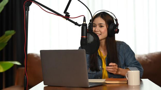 Stylish young woman wearing headphone using condenser microphone and laptop to recording podcast in home studio.