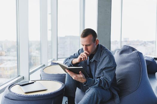 A male businessman in a casual suit resting in a comfortable pouf while working, an attractive freelancer in a suit works remotely and checks notes in a notebook