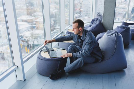 Break. Smiling young man relaxing on bean bag chair while sitting at table and relaxing using laptop, happy businessman leaning back, enjoying work, feeling satisfied