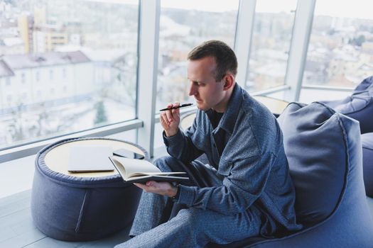 A young male student in a casual suit resting in a comfortable pouf while studying makes notes in a notebook, an attractive freelancer in a suit works remotely