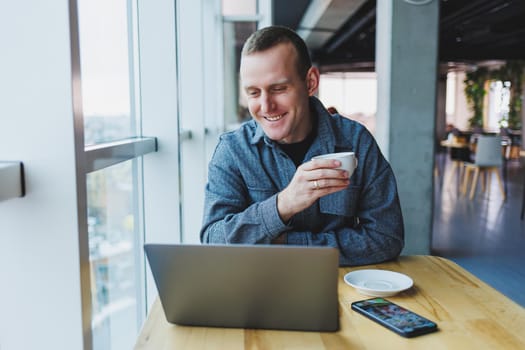 Successful happy business man is sitting at a table in a cafe, holding a cup of coffee and using a laptop.