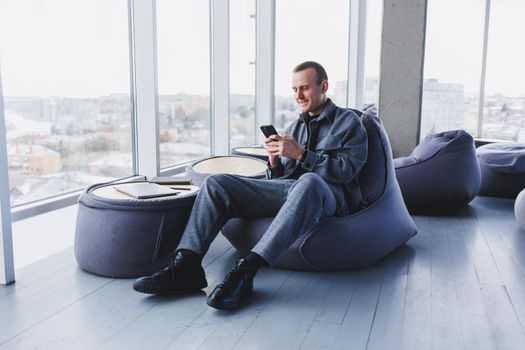 Young male student in a casual suit resting in a comfortable pouf while studying, an attractive freelancer in a suit works remotely